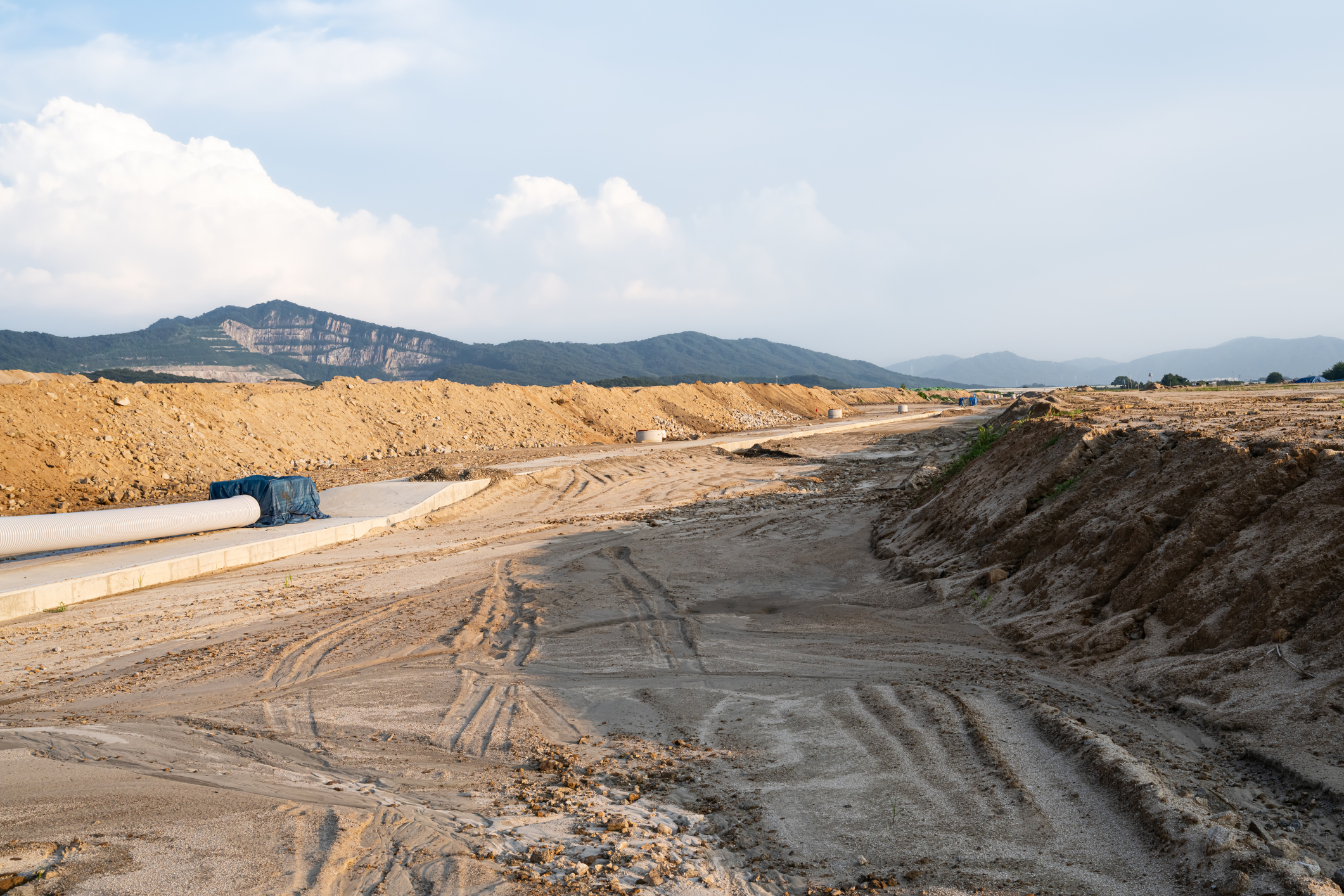 Expansive construction site with exposed earth and heavy machinery tracks. Surrounded by distant mountains, the scene captures early stages of land development and earthmoving operations.