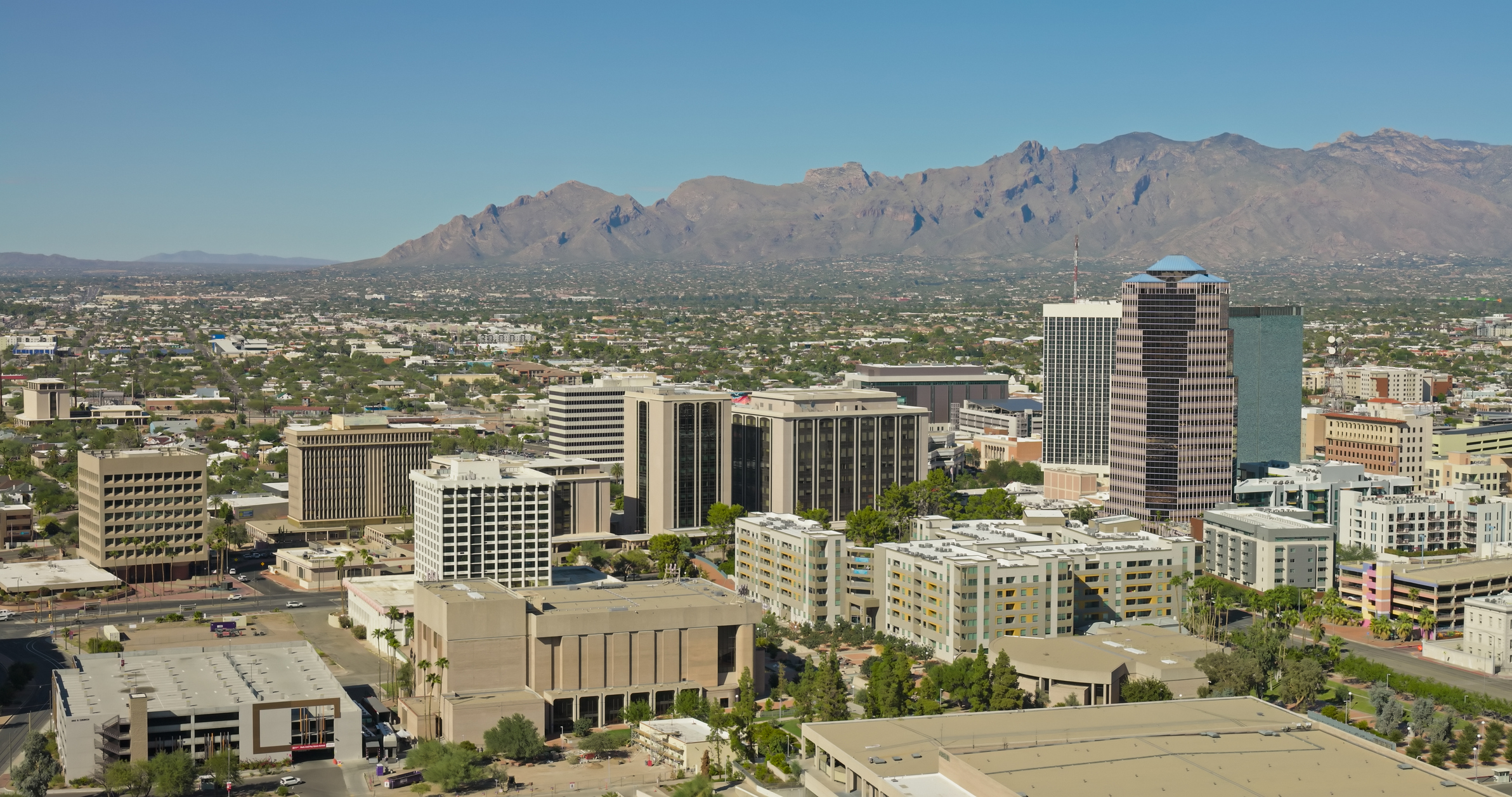 Aerial photograph of downtown Tucson, Arizona during the day with clear skies. The shot captures the central business district with its mix of modern high-rise buildings, office towers, and mid-rise structures set against the dramatic backdrop of the Santa Catalina Mountains. The urban core features contemporary glass and concrete architecture surrounded by low-rise residential neighborhoods that extend toward the desert foothills. The rugged mountain range creates a striking contrast with the developed cityscape below.